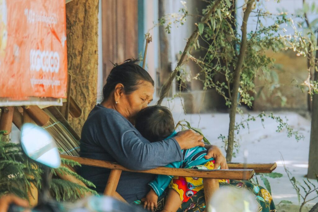 A Burmese grandmother lovingly embraces her grandchild in a peaceful outdoor setting.